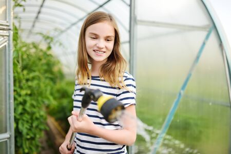 Adorable young girl watering variuos vegetables in a greenhouse on sunny summer day. Child helping with daily chores. Gardening activity for kids.の写真素材