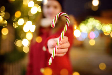 Cute young girl having striped candy cane on traditional Christmas fair in Riga, Latvia. Child enjoying sweets, candies and gingerbread on Xmas market. Winter time with family and kids.の写真素材