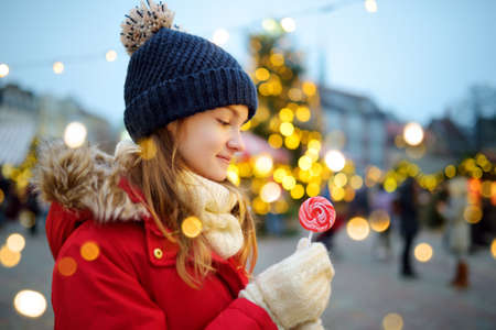 Cute young girl having striped lollipop on traditional Christmas fair in Riga, Latvia. Child enjoying sweets, candies and gingerbread on Xmas market. Winter time with family and kids.の写真素材