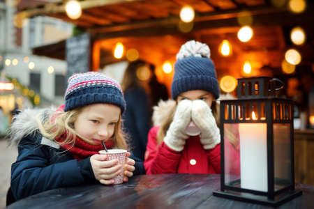 Two adorable sisters drinking hot chocolate on traditional Christmas fair in Riga, Latvia. Children enjoying sweets, candies and gingerbread on Xmas market. Winter time with family and kids.の写真素材