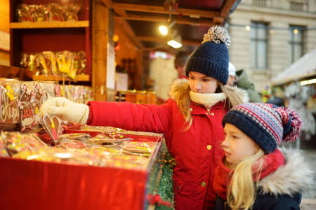 Cute young sisters choosing sweets on traditional Christmas market in Riga, Latvia. Kids buying candy and cookies on Xmas. Happy family time on chilly winter day.の写真素材