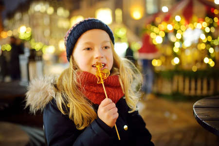 Cute young girl having rooster-shaped lollipop on traditional Christmas fair in Riga, Latvia. Child enjoying sweets, candies and gingerbread on Xmas market. Winter time with family and kids.の写真素材