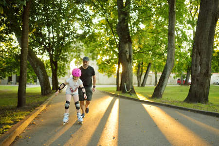 Father teaching his young daughter to roller skate on beautiful summer day in a park. Child wearing...