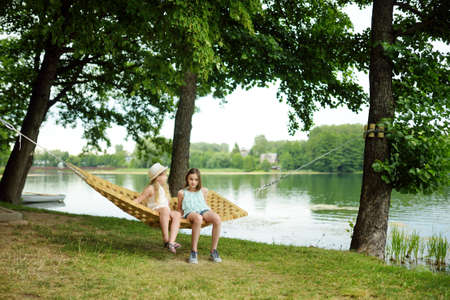 Cute young sisters having fun in hammock on beautiful summer day. Children relaxing in summer park. Family leisure at summer.の写真素材