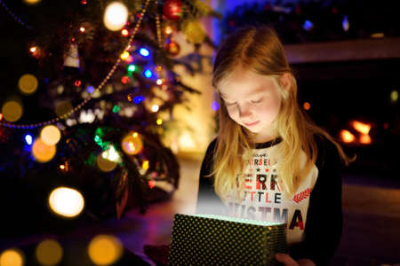Happy young girl wearing Christmas pajamas opening a gift box by a fireplace in a cozy dark living...