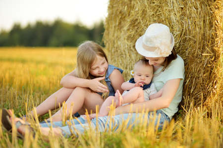 Big sisters and their baby brother having fun in a wheat field on a summer day. Children playing at hay bale field during harvest time. Kids enjoying warm sunset outdoors. Harvest in Lithuania.の写真素材