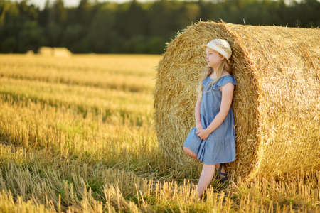Adorable young girl having fun in a wheat field on a summer day. Child playing at hay bale field during harvest time. Kid enjoying warm sunset outdoors. Harvesting crops in Lithuania.の写真素材