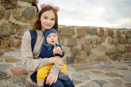 Cute teenage girl holding her baby brother while enjoying a view of Vilnius city from the Gediminas hill. Exploring tourist attractions with kids. Vilnius, Lithuaniaの写真素材