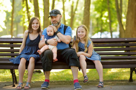 Father and three children having fun on summer day in city park. Adorable baby boy being held by his daddy. Two older sisters hugging their dad and baby brother. Big family. Kids with large age gap.の写真素材
