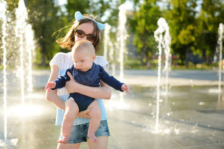 Cute big sister holding her baby brother by city fountain. Adorable teenage girl playing with her baby boy brother. Kids with large age gap. Children having fun with water on hot summer day.の写真素材
