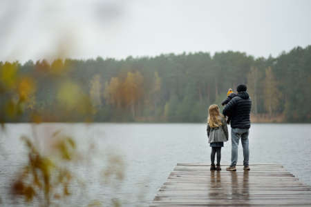 Father and two children having fun on late autumn day by the lake. Adorable baby boy being held by his daddy. Older sister hugging her dad and baby brother. Family exploring nature.の写真素材