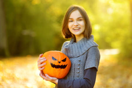 Cute young girl holding a small pumpkin with painted scary face on sunny autumn day. Kid trick or...
