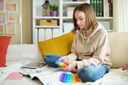 Teenage girl playing with rainbow pop-it fidget toy while studying at home. Teen kid with trendy stress and anxiety relief fidgeting game. Popping the dimples of sensory silicone toy.の写真素材