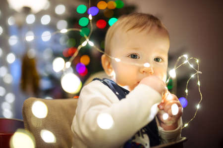 Happy little baby boy playing with Chistmas lights in a cozy living room on Christmas eve....