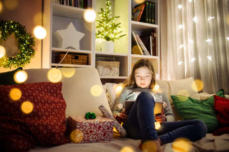 Cute young girl using a tablet pc at home in warm and cozy living room at Christmas. Family having online video call on Xmas eve. Winter evening at home with family and kids.の写真素材