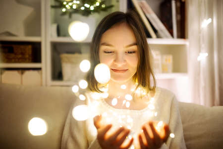 Beautiful teenage girl playing with Chistmas lights in a cozy living room on Christmas eve....