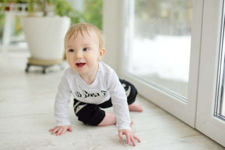 Adorable baby boy sitting by the window on sunny day. Toddler learning how to stand up unassisted.の写真素材