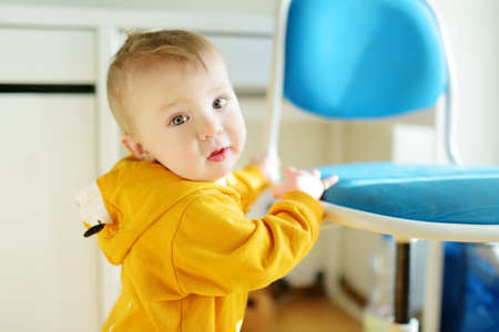 Adorable baby boy standing by the chair on bright living room. Toddler learning how to stand up...