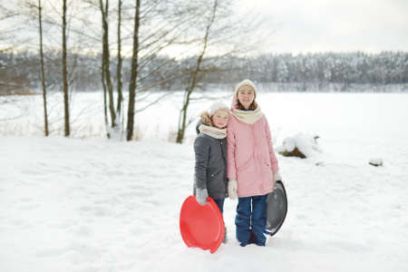 Two funny young girls having fun with a sleigh in beautiful winter park. Cute children playing in a snow. Winter activities for kids.の写真素材