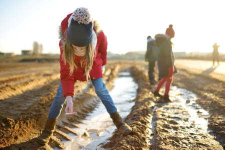 Group of children playing with thin ice puddles formed on the frozen soil in winter. Kids having fun in winter. Winter activities for kids.の写真素材