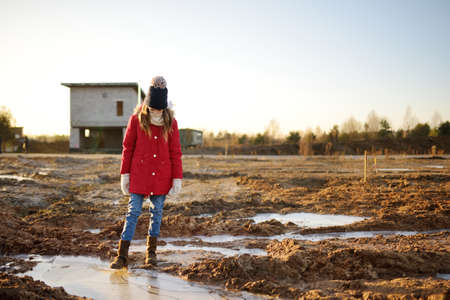 Happy young girl playing with thin ice puddles formed on the frozen soil in winter. Child having fun...