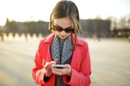 Pretty teenage girl using a smart phone in a city. Child browsing the Internet and posting online. Teenagers addiction to new technology.の写真素材