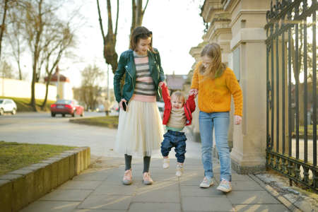 Two big sisters and their baby brother having fun outdoors. Two young girls holding their baby boy sibling on sunny spring day. Kids with large age gap. Kids during city walk.の写真素材