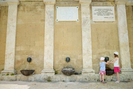 SORANO, ITALY - JUNE 8, 2019: Two sisters playing with a drinking water fountain in Sorano, an medieval hill town hanging from a tuff stone. Etruscan heritage. Province Grosseto, Tuscany, Italy.のeditorial素材