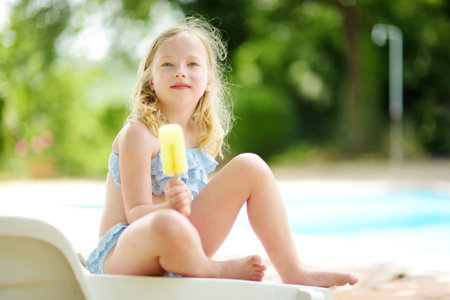 Cute young girl in a swimsuit having an ice cream by outdoor pool. Child having fun on hot summer day. Kid eating gelato on family vacations in Italy. Family activities by the water.の写真素材