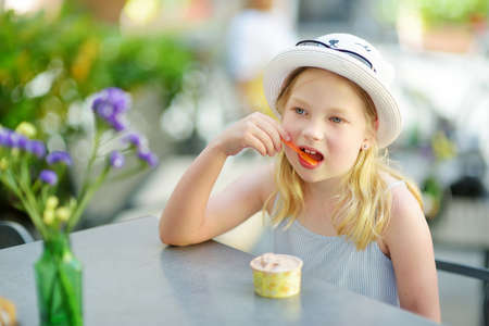 Adorable little girl in straw hat having an ice-cream on warm and sunny summer day during family vacations in Orvieto, Umbria Italy. Cute child sitting in outdoor cafe and eating a gelato.の写真素材