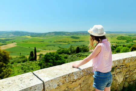 Young girl admiring the view of green fields and farmlands with small villages on the horizon. Summer rural landscape of rolling hills surrounding Pienza town, Tuscany, Italy.の写真素材