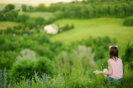Young girl admiring the view of green fields and farmlands with small villages on the horizon. Summer rural landscape of rolling hills surrounding Pienza town, Tuscany, Italy.の写真素材