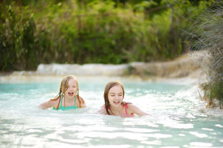 Two young sisters bathing in natural swimming pool in Bagno Vignoni, with thermal spring water and calcium carbonate deposits, which form white concretions and waterfall. Tuscany, Italy.の写真素材