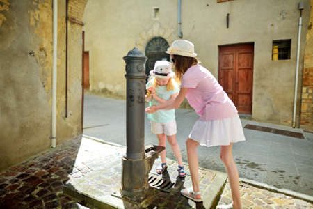 Two sisters playing with drinking water fountain in Montalcino town, located on top of a hill top and surrounded by vineyards, known worldwide for the production of delicious wine. Tuscany, Italy.の写真素材