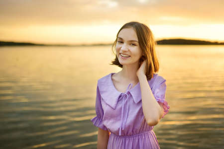 Beautiful teenage girl wearing pink dress having fun by a lake on warm and sunny summer day. Pretty young girl on a sunset. Summer activities for families.の写真素材