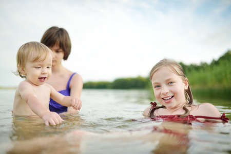 Cute toddler boy and his big teenage sisters playing by a river on hot summer day. Adorable child having fun outdoors during summer vacations. Water activities for kids.の写真素材