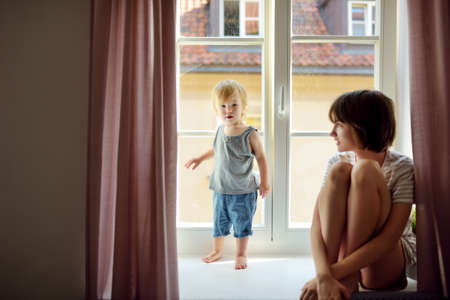 Teenage sister and her toddler brother looking out the window. Siblings sitting on a windowsill....