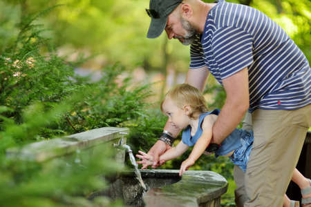 Father helping his toddler son to wash his hand in city fountain. Dad and son having fun on sunny summer day in the city.の写真素材