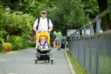Young father walking in a park with toddler son in pushchair. Man pushing a stroller for toddler...