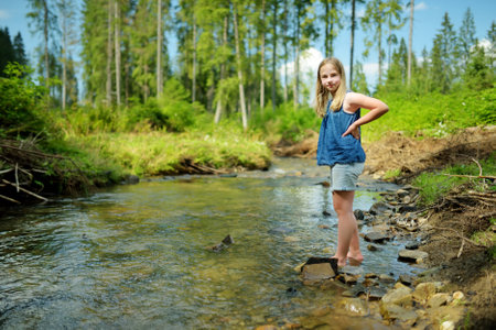 Young girl near cold shallow stream winding through majestic pine trees of Tatra mountain range near Zakopane, Poland. Beautiful Low Tatras alpine landscape with forest in the foregroundの写真素材