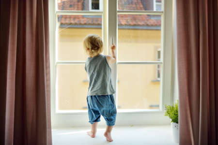 Cute toddler boy standing on a windowsill. Adorable child looking out the window. Window with lock...