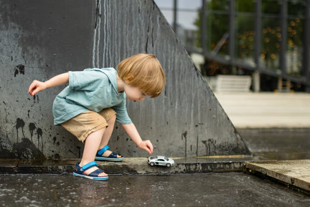 Cute toddler boy playing with yellow toy car outdoors. Kid having fun with water in city fountain. Summer activities for little children.の写真素材