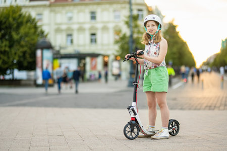 Adorable young girl riding her scooter in a city on sunny summer evening. Pretty preteen child riding a roller. Active leisure and outdoor sports for kids.の写真素材