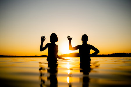 Silhouettes of teenage girls having fun by a lake on warm and sunny summer evening. Pretty young girls on a sunset. Summer activities for families.の写真素材
