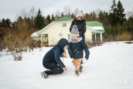 Adorable toddler boy and his older sisters having fun in a backyard on snowy winter day. Cute children wearing warm clothes playing in a snow. Winter activities for family with kids.の写真素材
