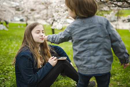 Pretty teenage girl and her toddler brother having fun in blooming cherry tree garden on beautiful...
