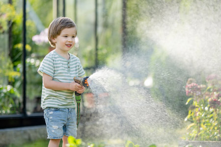 Cute little boy watering flower beds in the garden at summer day. Child using garden hose to water vegetables. Kid helping with everyday chores. Mommy's little helper.の写真素材