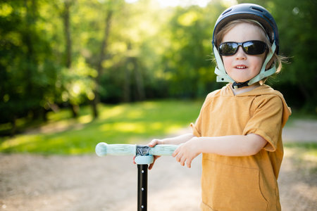 Adorable little boy riding his scooter in a city park on sunny summer evening. Young child riding a roller with a helmet on. Active leisure and outdoor sports for kids.の写真素材