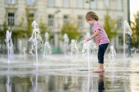 Cute little boy having fun with water in city fountain. Child playing water games outdoors on hot day. Summer activities for small children.の写真素材