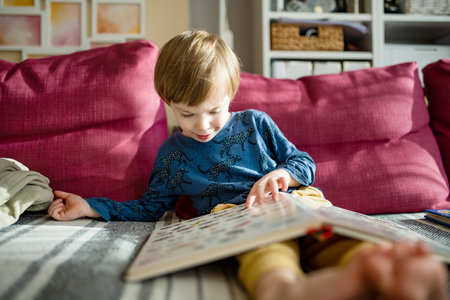 Cute little boy reading books on the sofa at home on a bright sunny afternoon. Daytime care creative...